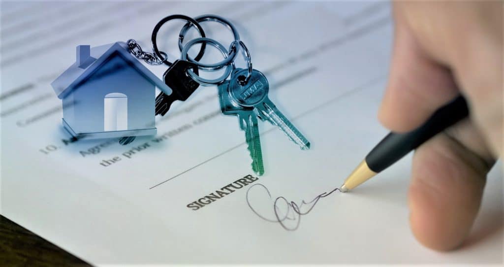 Someone signing a contract with a set of house keys on top of the paper, the keychain in a miniature house