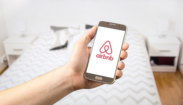 Person standing in a white bedroom holding a cellphone showing the Airbnb logo.