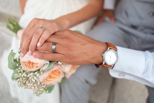 Closeup of the hands of the bride and groom, showing their rings and bouquet of roses